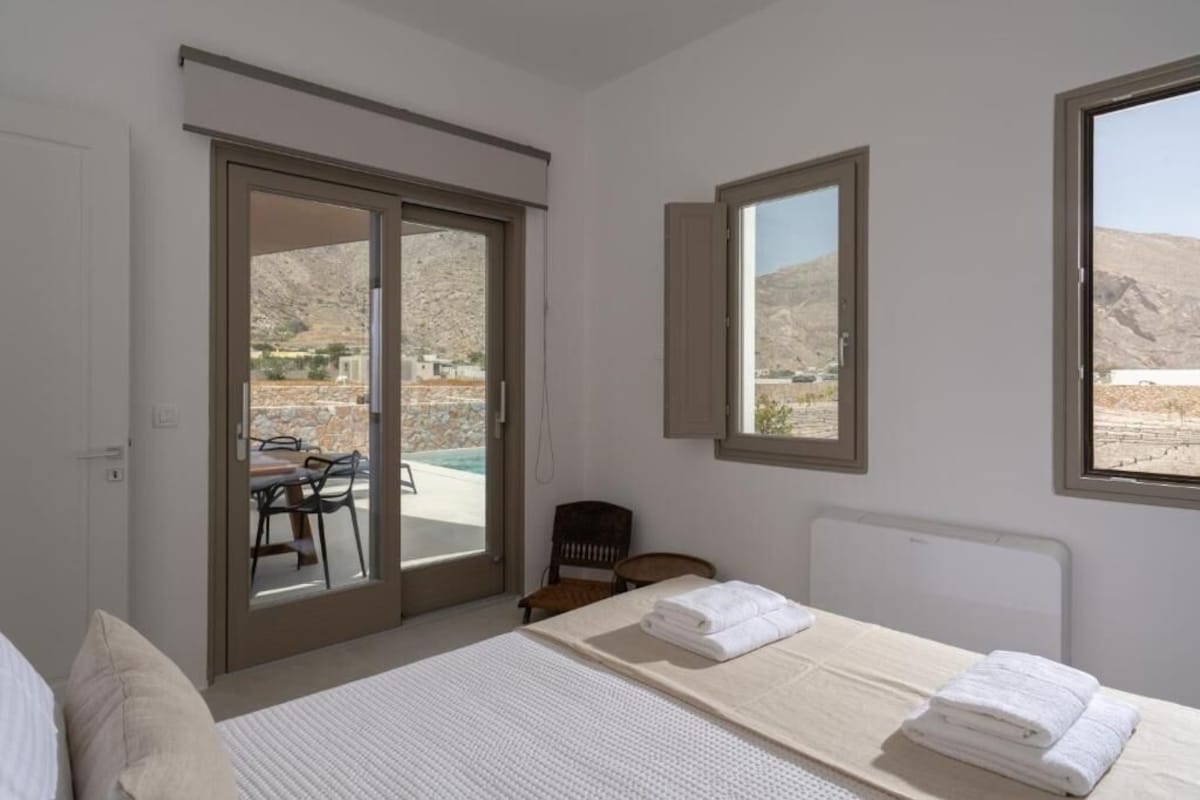 A bedroom with a bed, a chair, a table, and a view of a pool and mountains.
