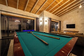 Une table de billard avec des queues de billard, un canapé et un téléviseur, dans une pièce aux murs en pierre et aux poutres apparentes.