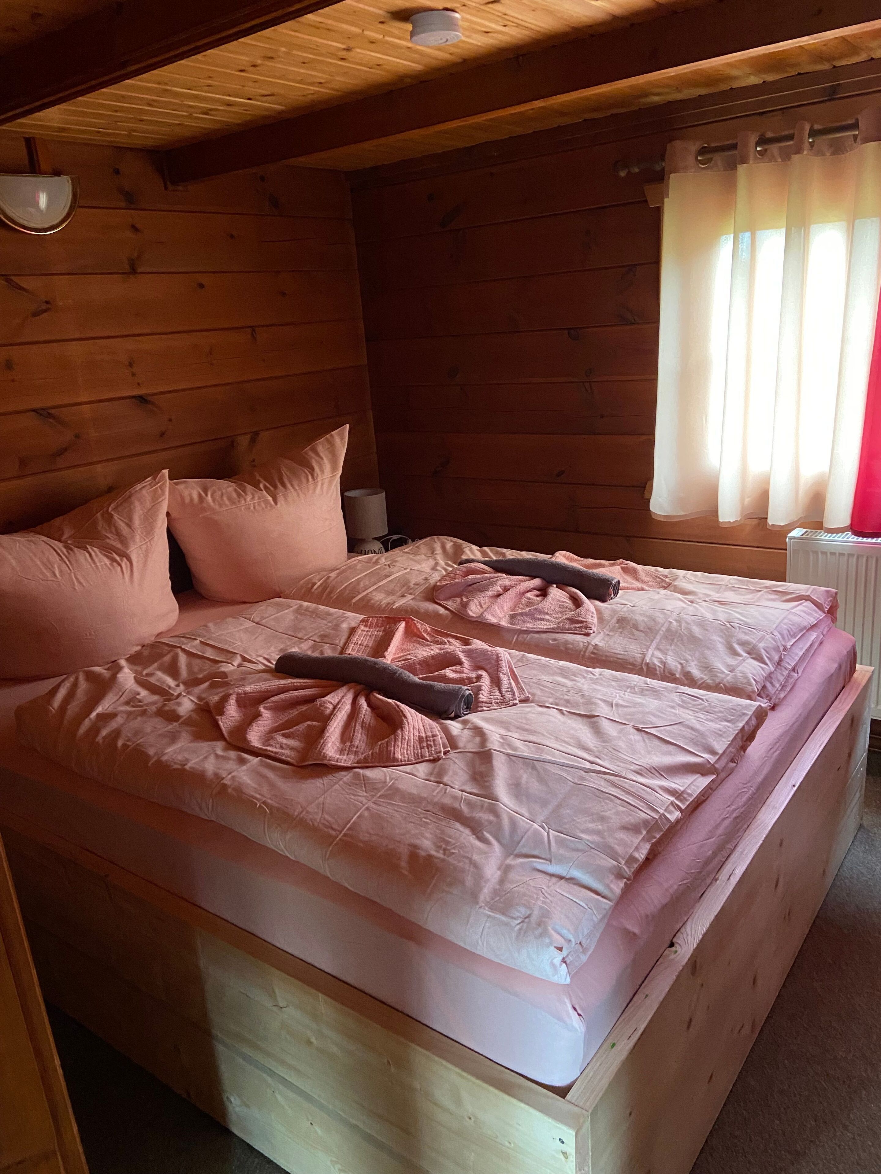 A wooden cabin bedroom with two beds, a window with curtains, and a ceiling light.