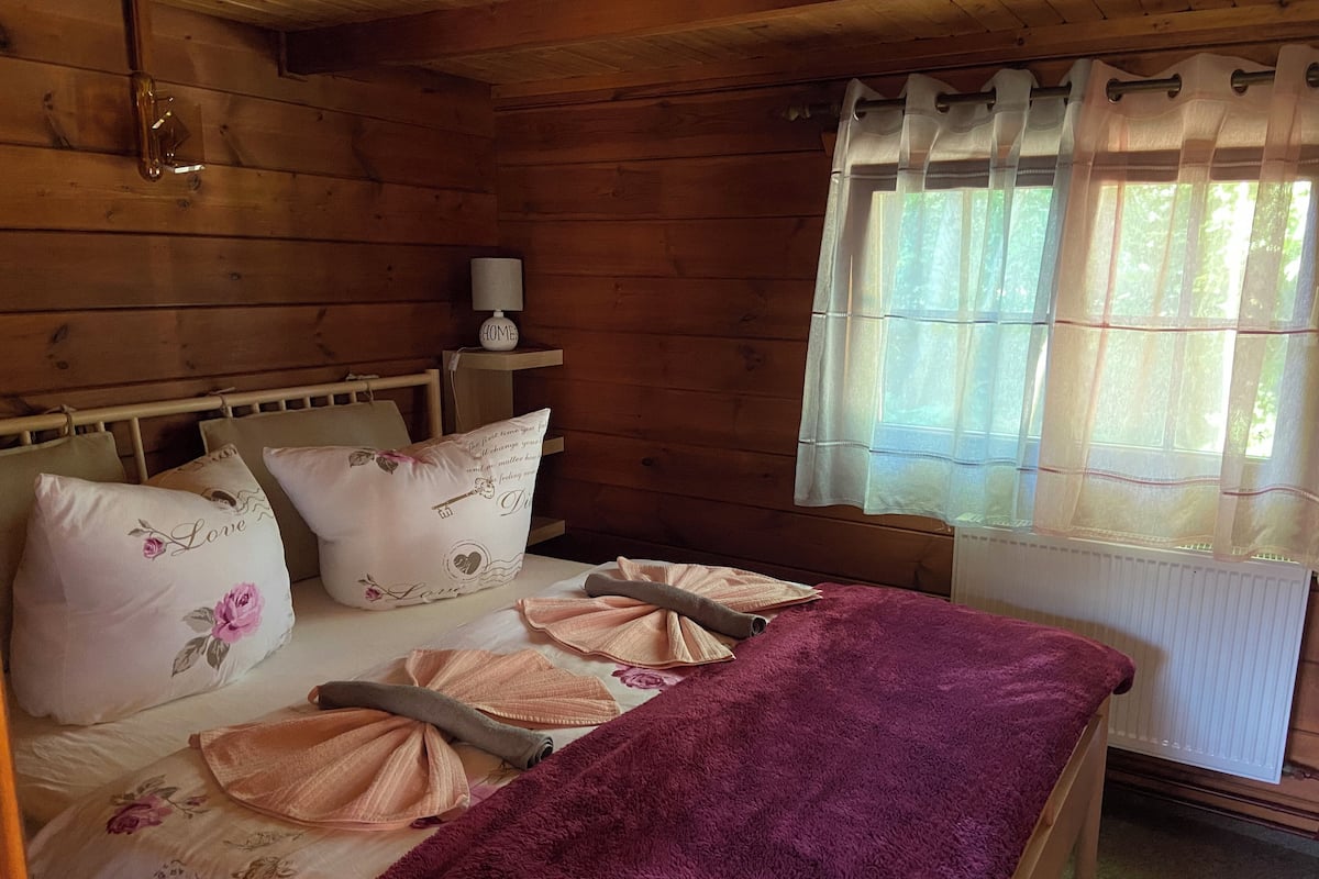 Une chambre à coucher dans une cabane en bois, avec un lit, des oreillers, une table de chevet, une lampe et une fenêtre avec des rideaux.
