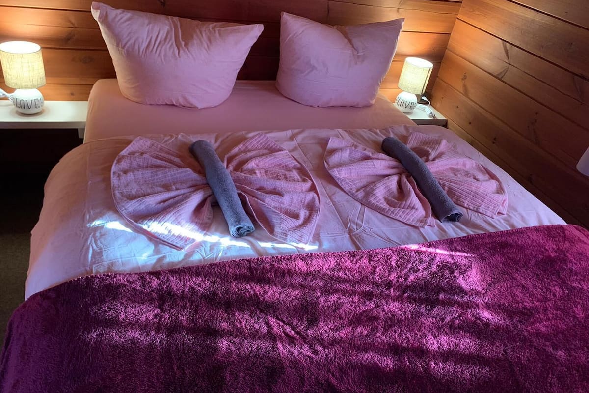 Un lit bien fait, avec du linge de lit rose et deux serviettes enroulées, une tête de lit en bois et des lampes de chevet.