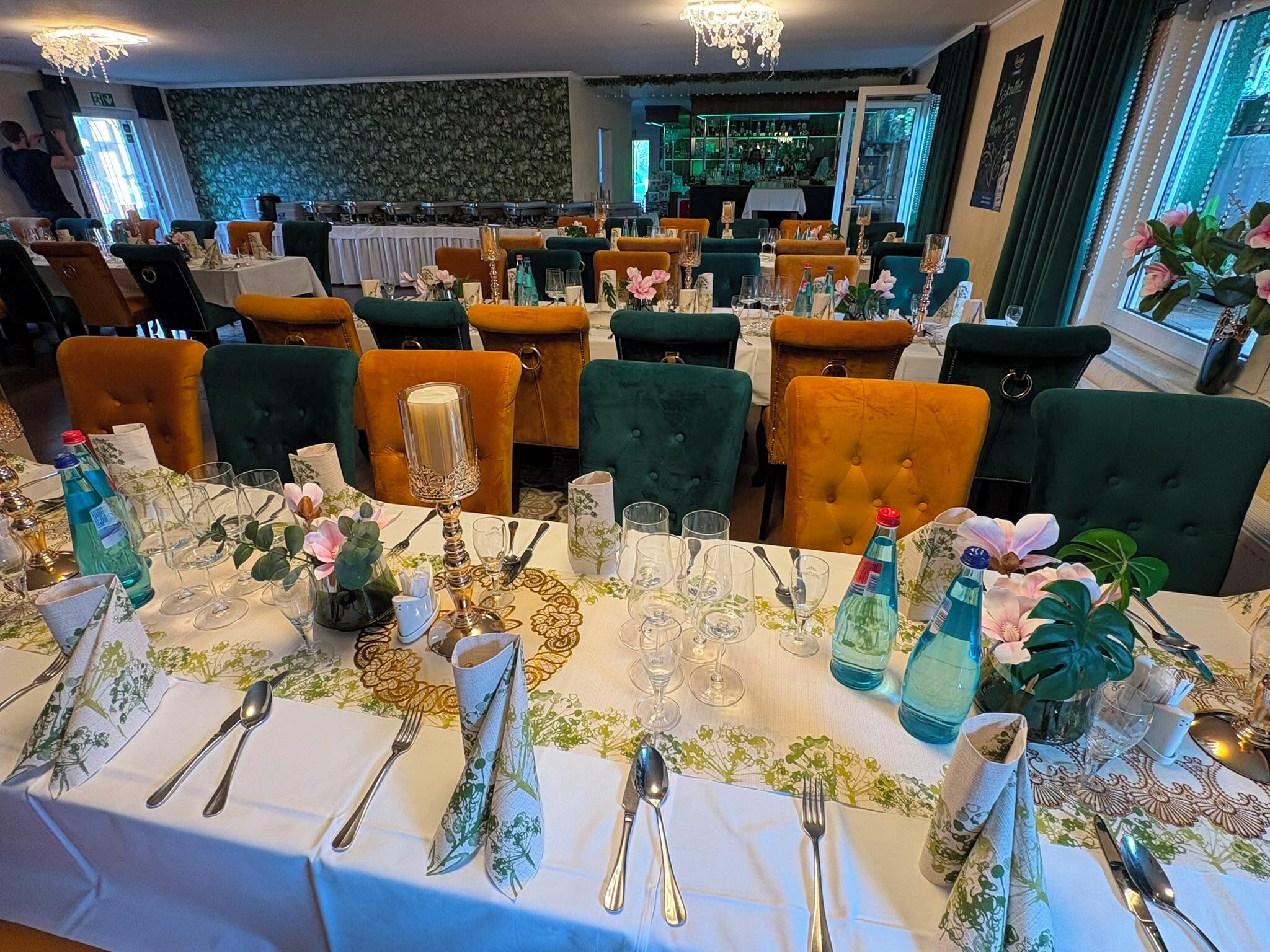 Un restaurante con varias mesas dispuestas para comer, con sillas tapizadas en verde y naranja, manteles blancos y centros de mesa florales.