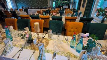 Un restaurante con varias mesas dispuestas para comer, con sillas tapizadas en verde y naranja, manteles blancos y centros de mesa florales.