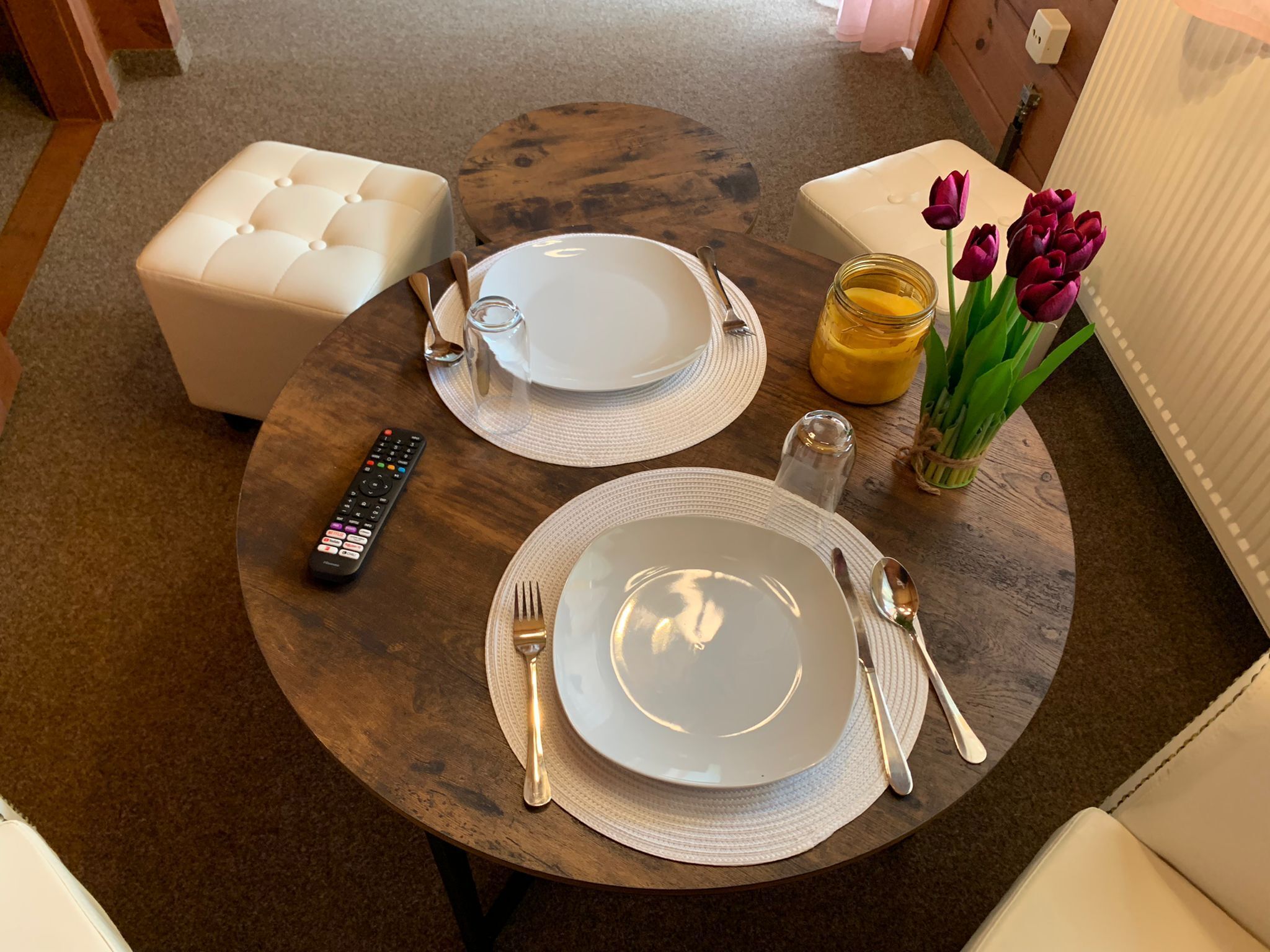Une table à manger dressée avec des assiettes, des couverts, une télécommande, un salier, un pot de confiture et des tulipes.