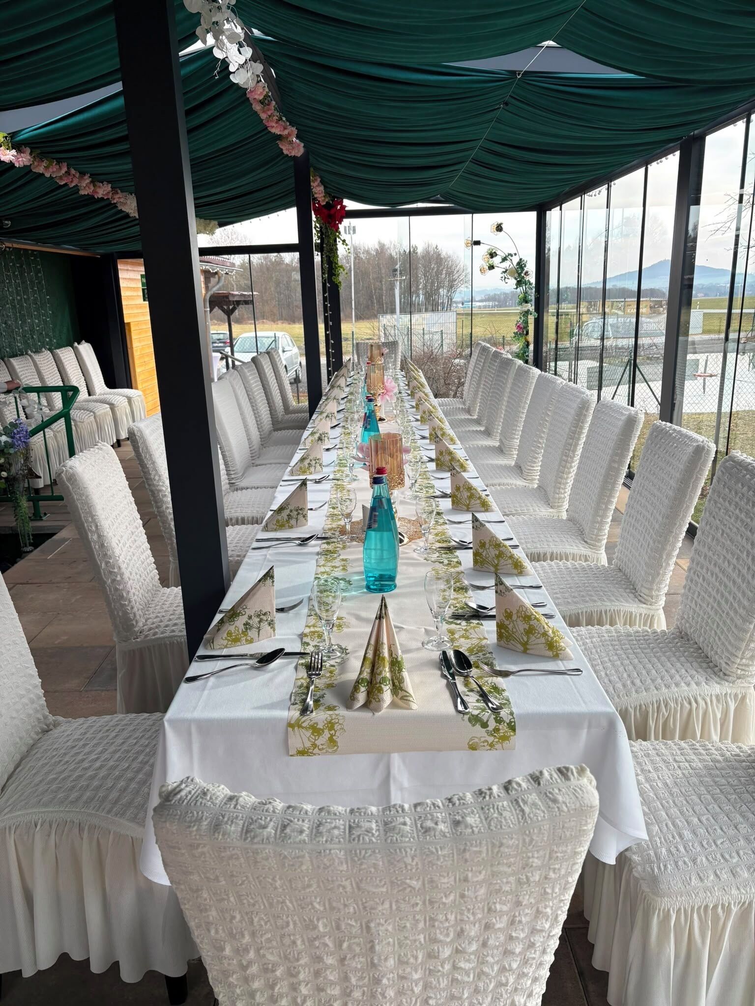 Una larga mesa de comedor dispuesta con sillas blancas, una botella color turquesa y centros de mesa florales.