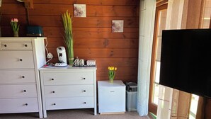 Ein kleiner, gemütlicher Raum mit Holzwänden, einem weißen Schminktisch, einem kleinen Kühlschrank, einem Fernseher und einem Fenster mit Vorhängen.
