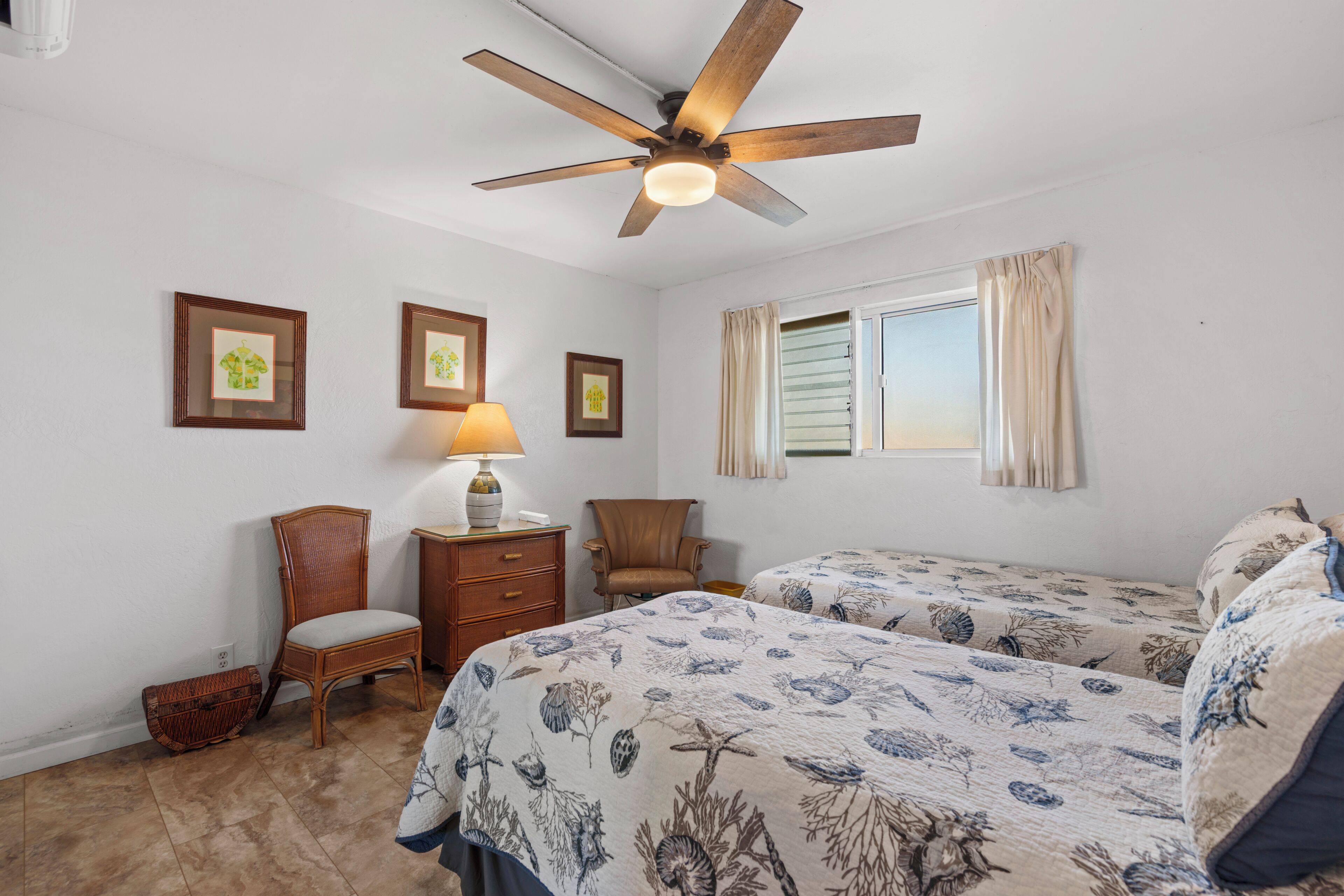 A bedroom with two beds, a ceiling fan, a window with curtains, and framed pictures on the wall.