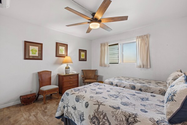 A bedroom with two beds, a ceiling fan, a window with curtains, and framed pictures on the wall. - Beautiful Ocean View 2BR w/ Split AC & Private Lanai – Menehune Shores #524 (Kihei, Maui)