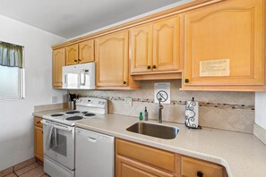 A kitchen with wooden cabinets, a white stove, a sink, and a tiled backsplash. - Beautiful Ocean View 2BR w/ Split AC & Private Lanai – Menehune Shores #524 (Kihei, Maui)