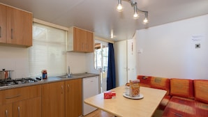 Eine kleine Küche mit Holzschränken, einem Herd und einem Spülbecken. Ein Esstisch steht dort, mit einer Schachtel Frühstücksflocken und einer Tasse darauf. Ein kleiner Kühlschrank ist sichtbar, und das Zimmer hat ein Fenster mit Jalousien.