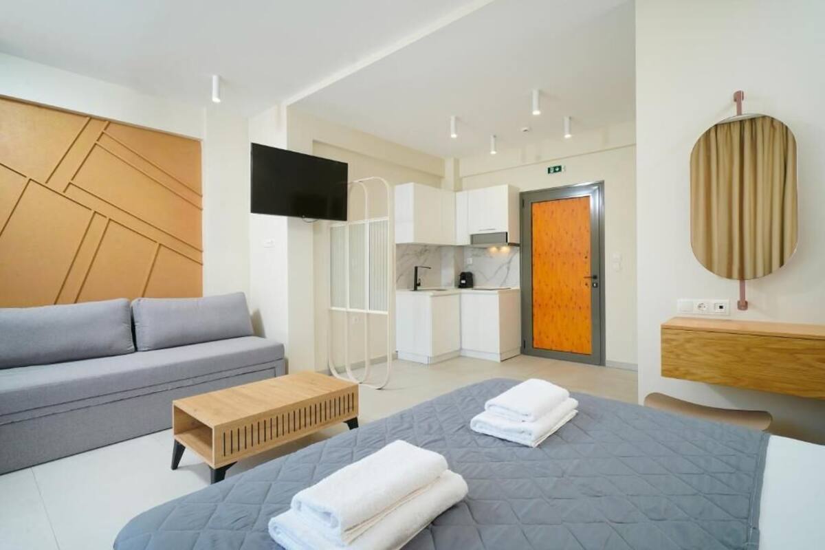A modern hotel room with a grey sofa, a bed with folded towels, a wooden coffee table, a TV mounted on the wall, a kitchen area, and a wooden panel wall.