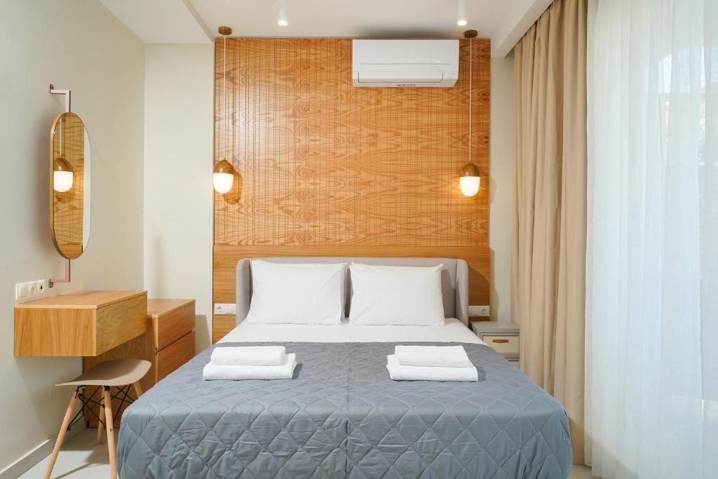 A hotel room with a wooden headboard, a bed with two pillows, a bedside table with a lamp, a mirror, and a desk with drawers.
