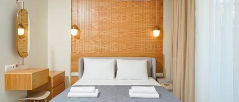A hotel room with a wooden headboard, a bed with two pillows, a bedside table with a lamp, a mirror, and a desk with drawers.
