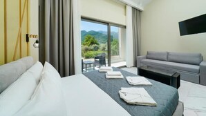 A hotel room with a bed, a sofa, a flat-screen TV, and a balcony with a view of the mountains. - Valen Mare Suites (Thasos)