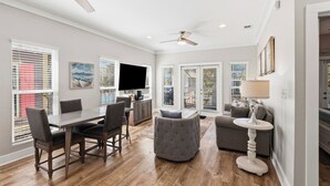 A modern living room with a dining area, large windows, a flat-screen TV, and a ceiling fan. - Seacrest 3BR Cottage at Camp Creek – Pool, Hot tub  & Beach Nearby! (Seacrest)