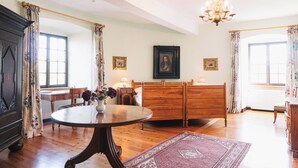 A room with a wooden bed, a round table with a vase of flowers, a chandelier, and two windows with floral curtains. - Gemünden Castle with pool. Paradise for hikers! (Gemünden)