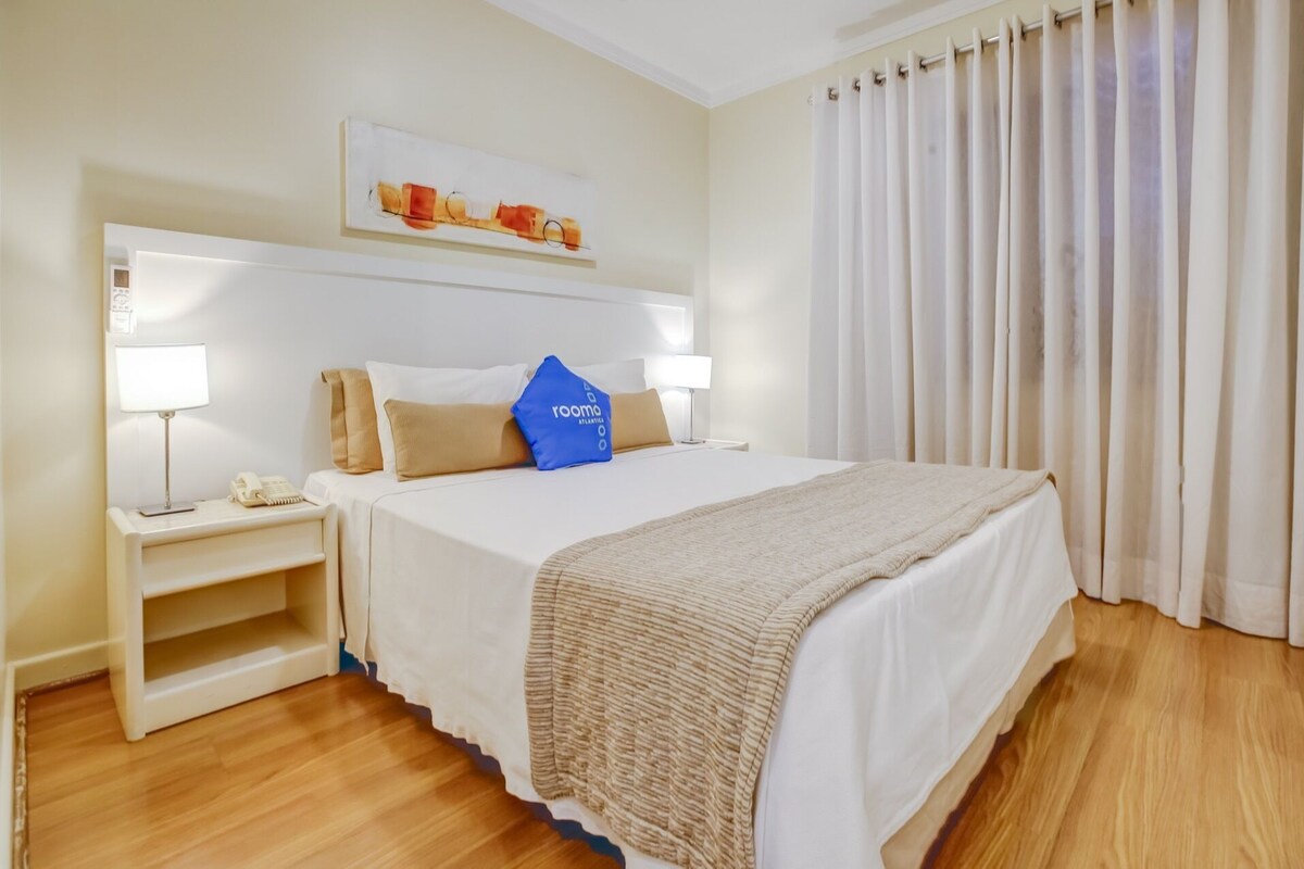 Una habitación de hotel con una cama, mesitas de noche, una lámpara, un teléfono, una almohada azul y un cuadro en la pared.