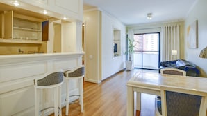 A modern kitchen with white cabinetry, a dining area with a table and two bar stools, and a living room with a sofa and a large window. - 360 Itaim Bibi (São Paulo)