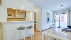 A modern kitchen with white cabinetry, a microwave, and two bar stools. There is a dining table with a blue accent and a potted plant by the window. - 360 Itaim Bibi (São Paulo)
