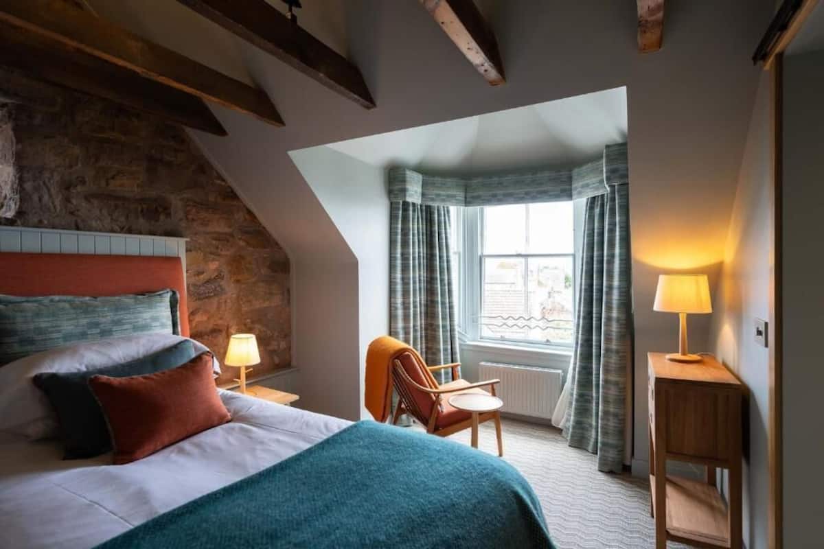 Un dormitorio con cama, una silla, una mesa y una ventana con cortinas.