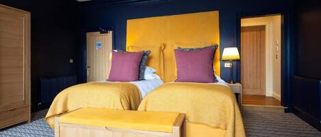 A hotel room with two beds, a wooden bench, a wardrobe, and a door leading to another room.