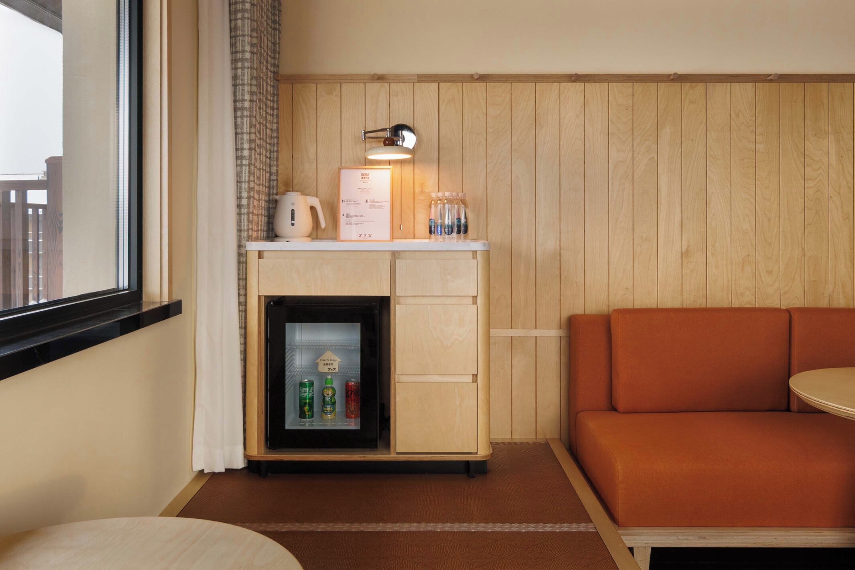 Ett litet rum med en väggpanel i trä, ett litet pentry med kylskåp, en kaffebryggare och en flaska vatten. Det finns en rund, orange soffa och ett litet runt bord.