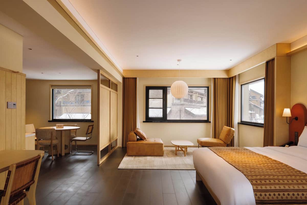 Ein Hotelzimmer mit einem großen Bett, einer Couch, einem kleinen Tisch und einem Sessel. Zwei Fenster mit Vorhängen, eine Hängeleuchte und ein Holzboden sind ebenfalls vorhanden.