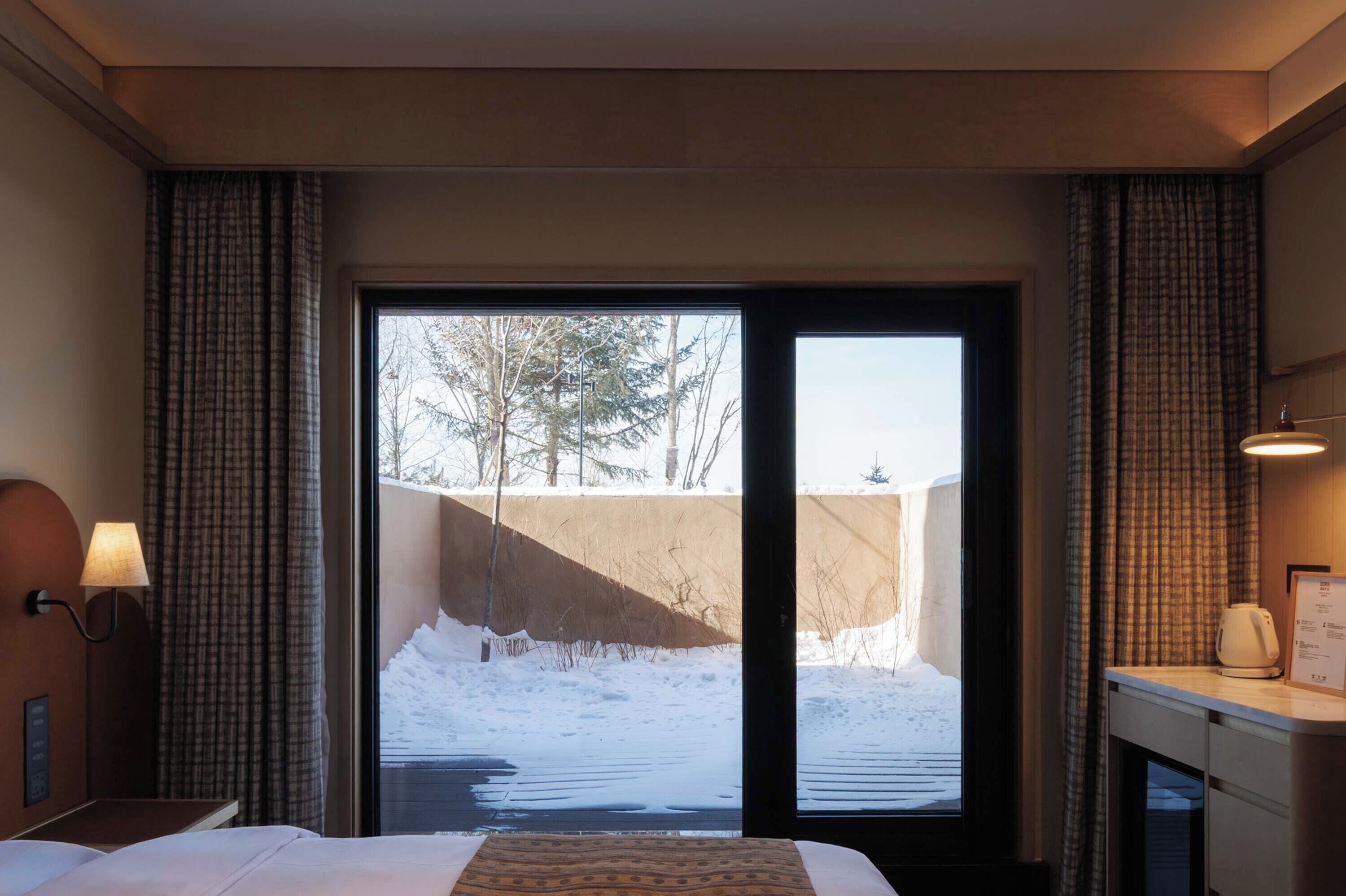 A hotel room with a bed, a desk with a lamp, and a view of a snowy outdoor area through a sliding door.