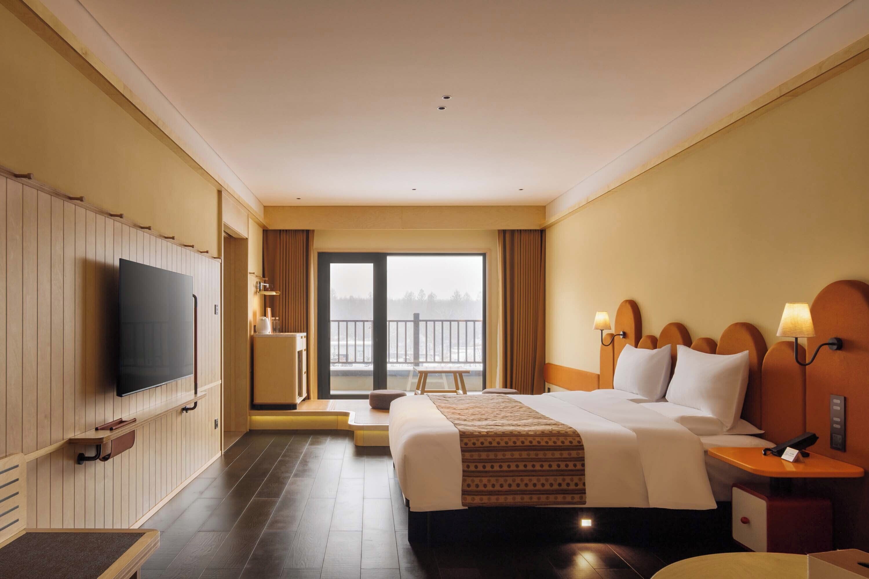 Habitación de hotel con una cama grande, televisión, escritorio con lámpara, una silla y vistas a un balcón.