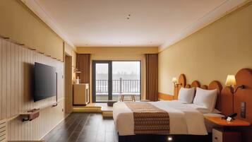 Habitación de hotel con una cama grande, televisión, escritorio con lámpara, una silla y vistas a un balcón.