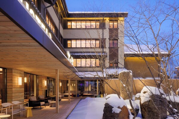 Un hotel moderno con una zona de estar exterior cubierta, ventanas amplias y rocas cubiertas de nieve.