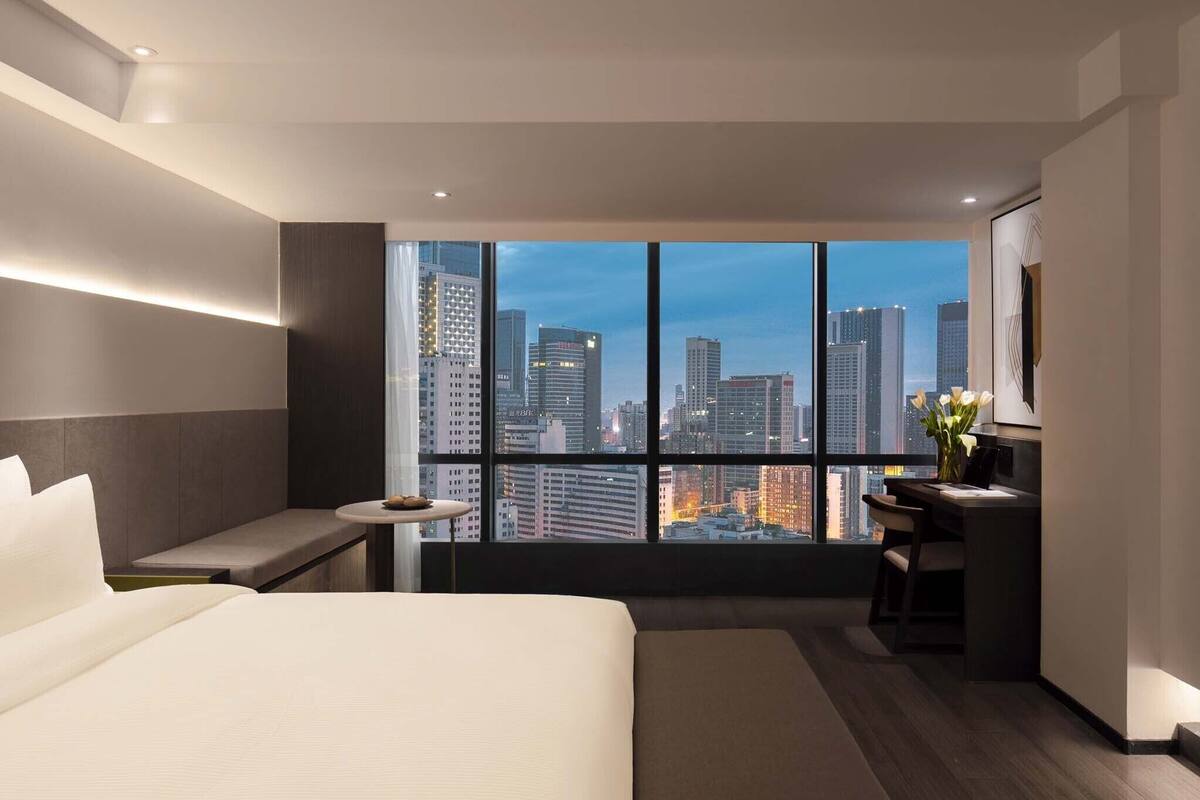 Una habitación de hotel moderna con un ventanal amplio que ofrece vistas a la ciudad, una cama, un escritorio y un pequeño rincón de descanso.