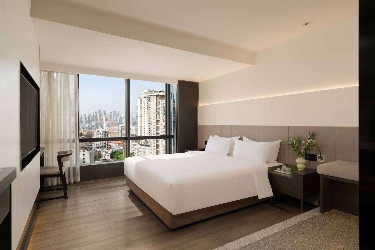 Una habitación de hotel moderna con una cama grande, una mesita de noche con un jarrón de flores, una silla y vistas de la silueta urbana a través de amplios ventanales.