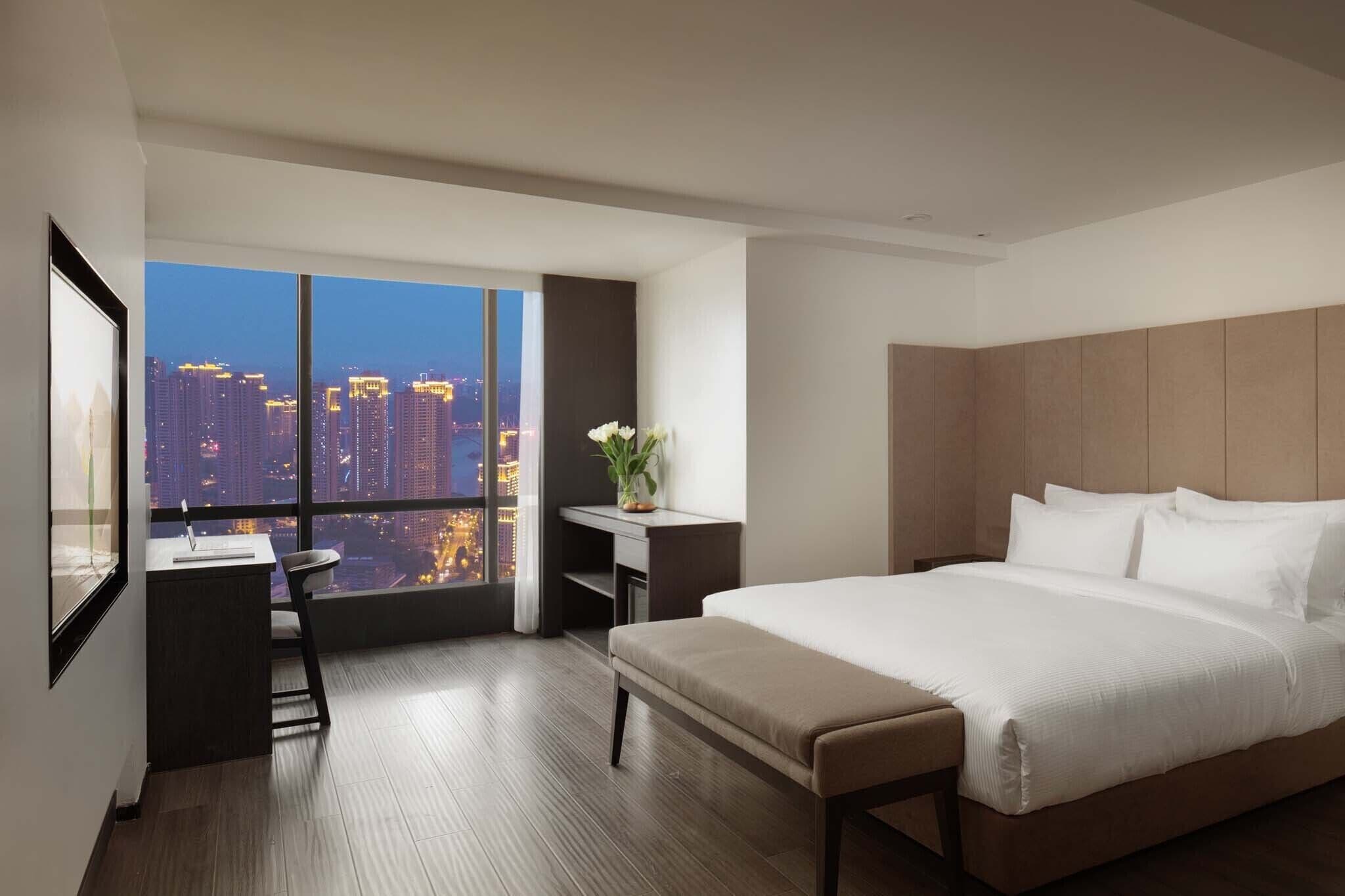 A modern hotel room with a large bed, a desk, and a view of a cityscape at dusk.
