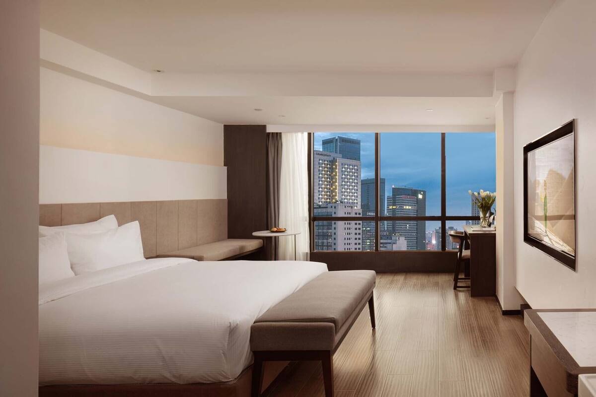 Una habitación de hotel moderna con una cama grande, un escritorio y vistas de la ciudad a través de amplias ventanas.