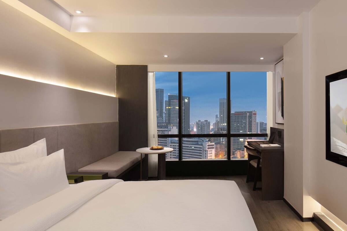 Habitación de hotel moderna con un ventanal grande que ofrece vistas a la ciudad, una cama, un escritorio y un pequeño rincón de descanso.