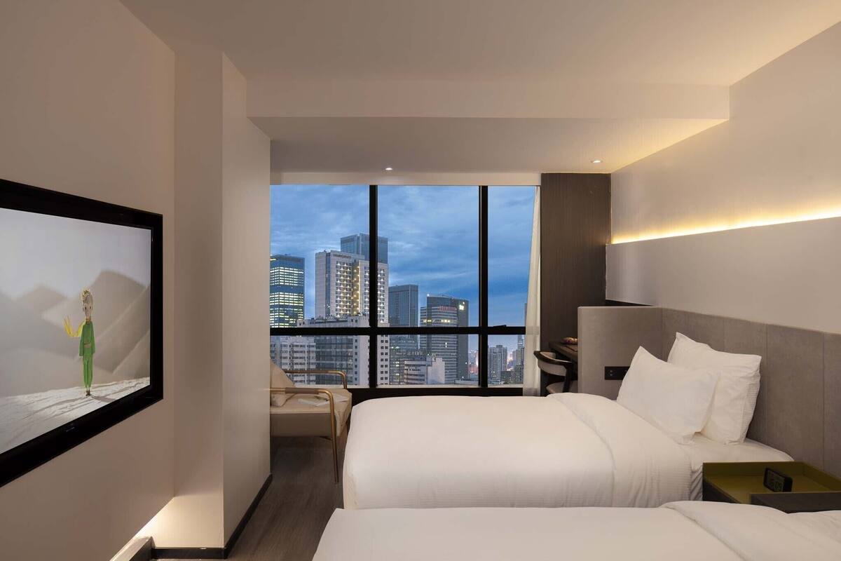 Habitación de hotel moderna con un amplio ventanal que ofrece vistas del horizonte urbano, una cama con ropa blanca, una mesita de noche y una silla.