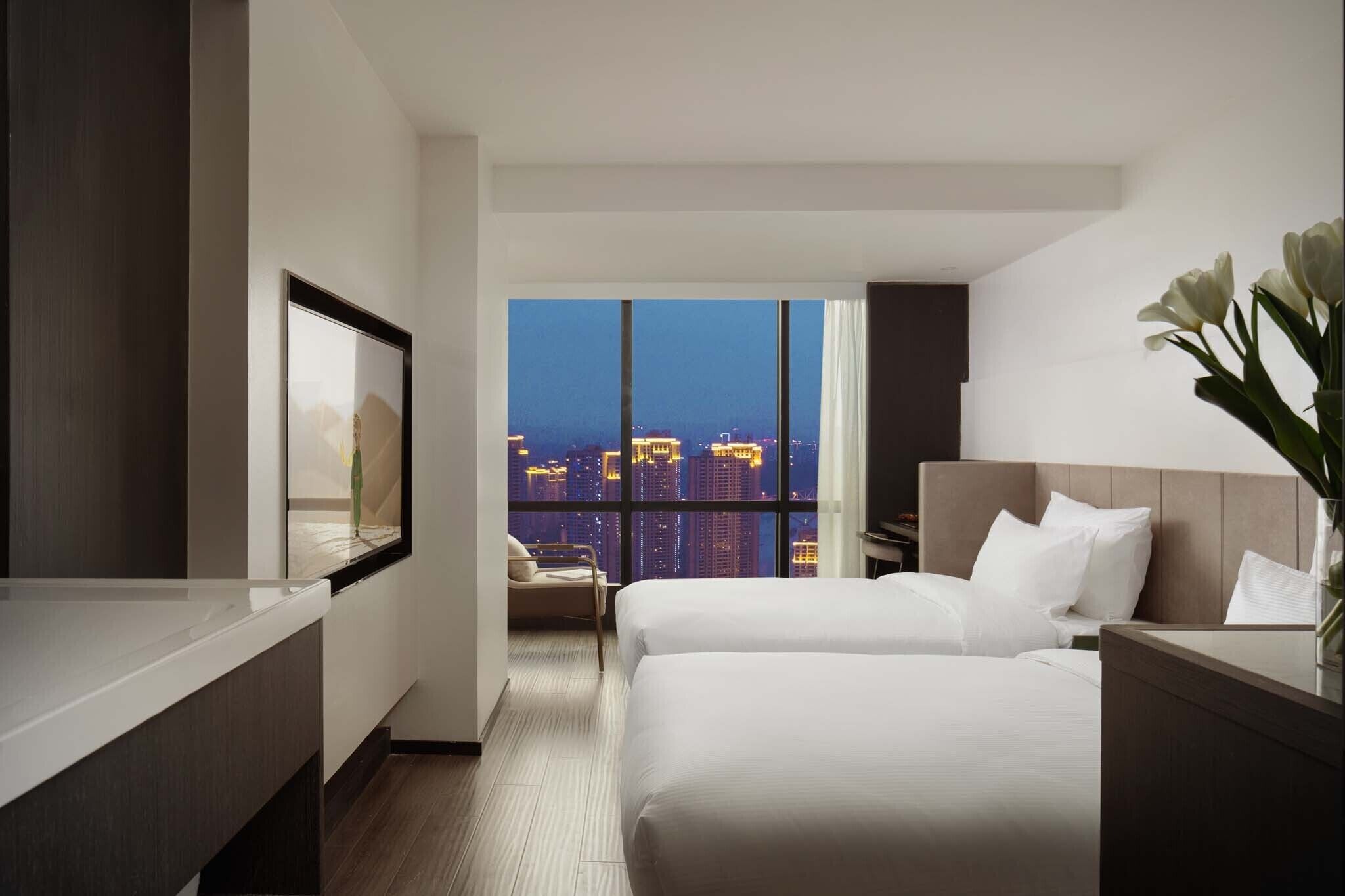 Habitación de hotel con dos camas, un ventanal grande con vista a la ciudad, un escritorio y una silla.