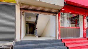 Ein Gebäudeeingang mit einer Treppe, die zu einer Tür führt, einem geschlossenen Rollo links und einer roten Markise rechts.