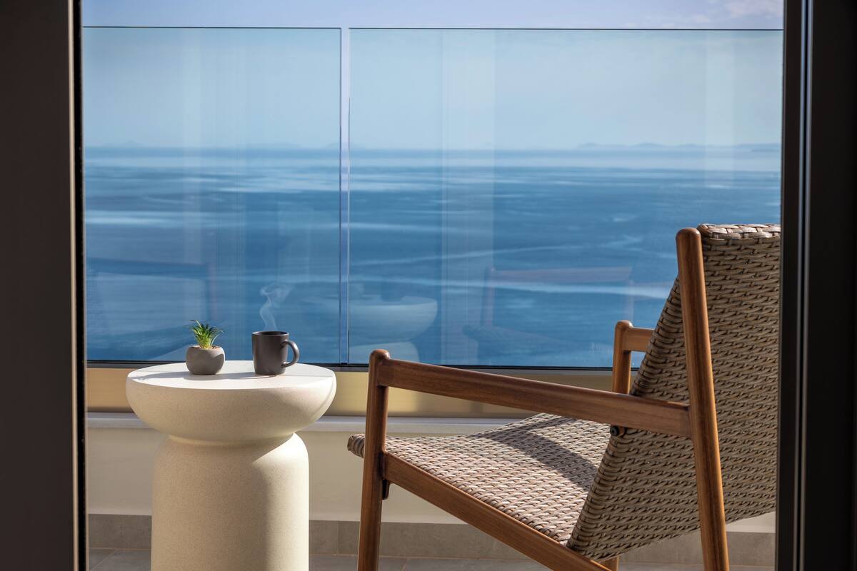 Un balcon avec vue sur l’océan, une chaise en osier, une petite table avec une plante et une tasse.