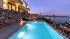 A modern poolside area with a stone wall, lounge chairs, and a view of the sea. - Elounda Infinity Blue Luxury Villas (Agios Nikolaos)