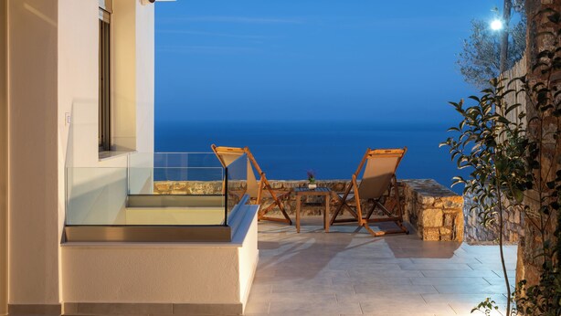 A terrace with a glass balustrade, two wooden chairs, a small table, and a view of the sea at dusk. - Elounda Infinity Blue Luxury Villas (Agios Nikolaos)