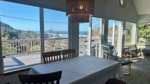 Ein Essbereich mit Tisch und Stühlen, große Fenster mit Blick auf das Meer und die Hügel sowie eine gewebte Pendelleuchte.