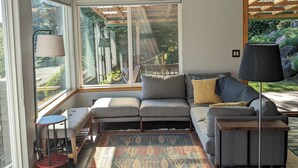 Ein modernes Wohnzimmer mit einem grauen Ecksofa, einem gemusterten Teppich und einem großen Fenster mit Blick auf einen Garten.