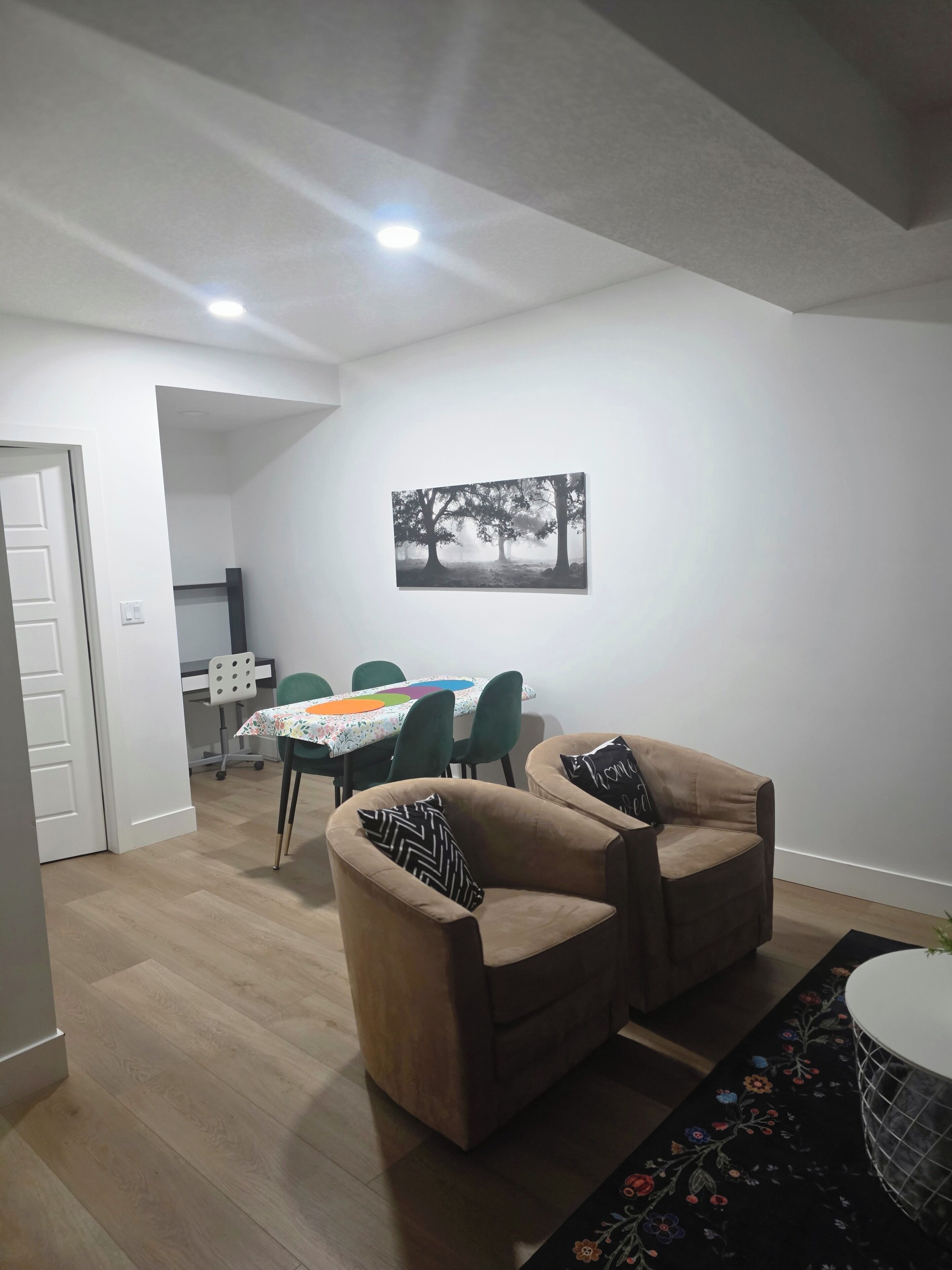 Ein modernes Wohnzimmer mit zwei beigen Sesseln, einem Esstisch mit bunten Tischdecken und einem gerahmten Bild an der Wand.