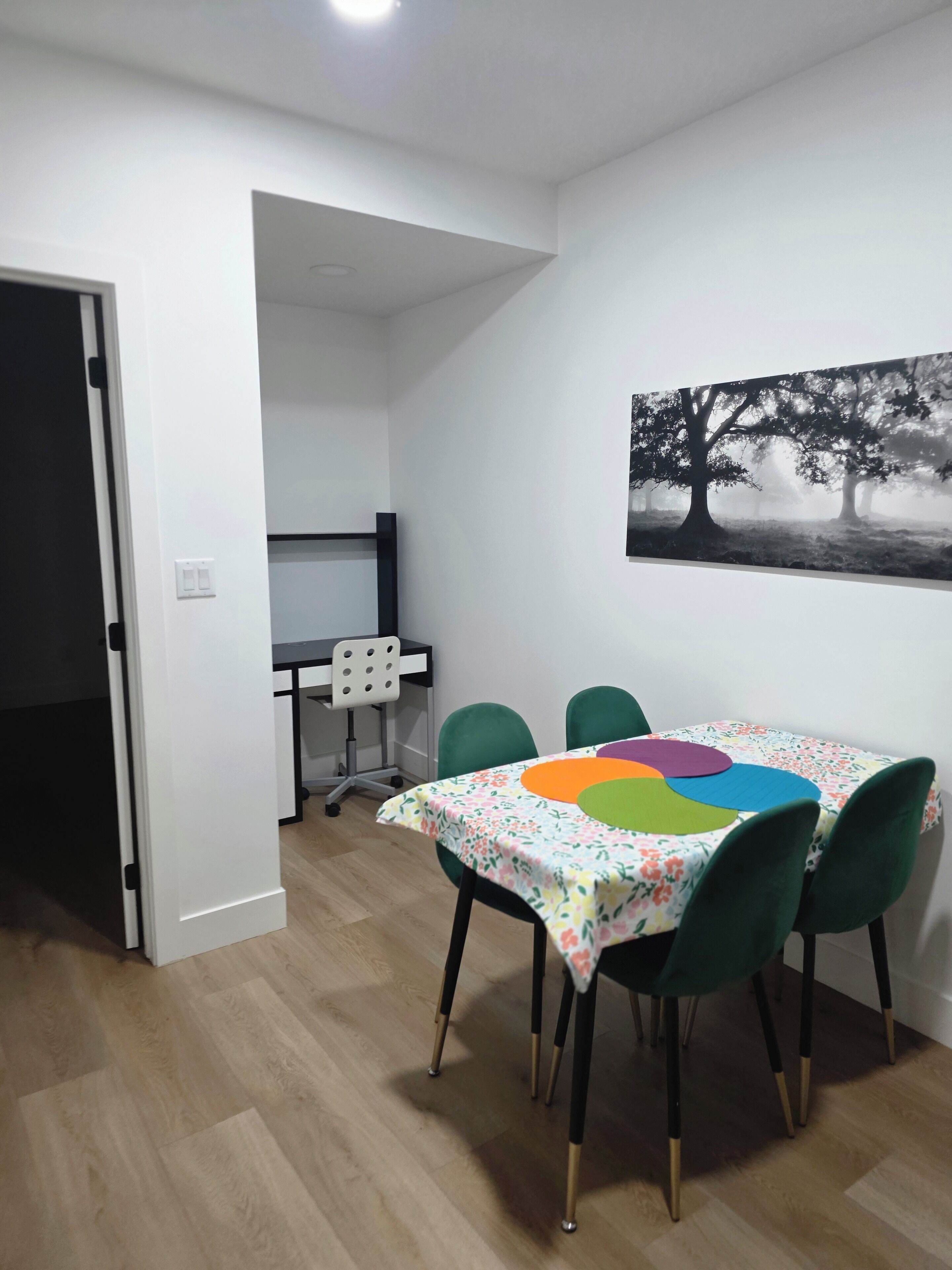 Een eetruimte met een tafel bedekt met een bloemig tafelkleed, groene stoelen en een schilderij van bomen aan de muur.