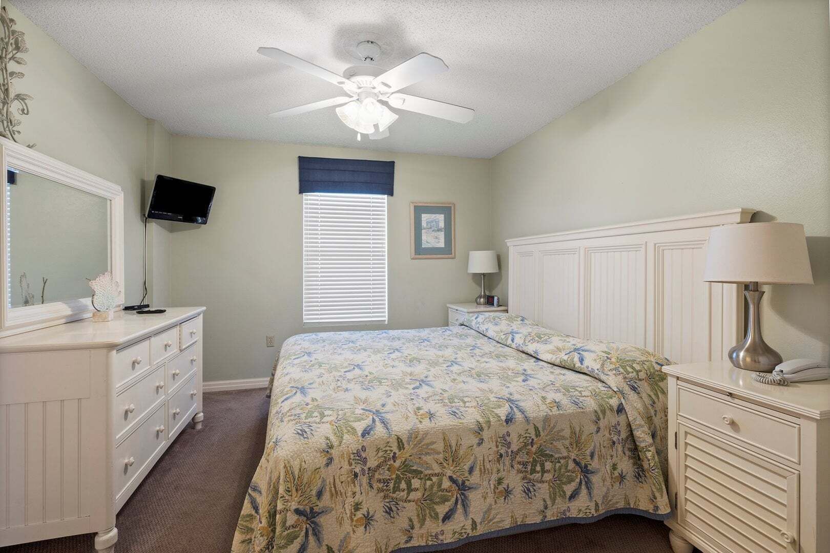 Ein Schlafzimmer mit einem Bett, einem Schminktisch, einem Nachttisch, einer Lampe, einem Fernseher und einem Deckenventilator.