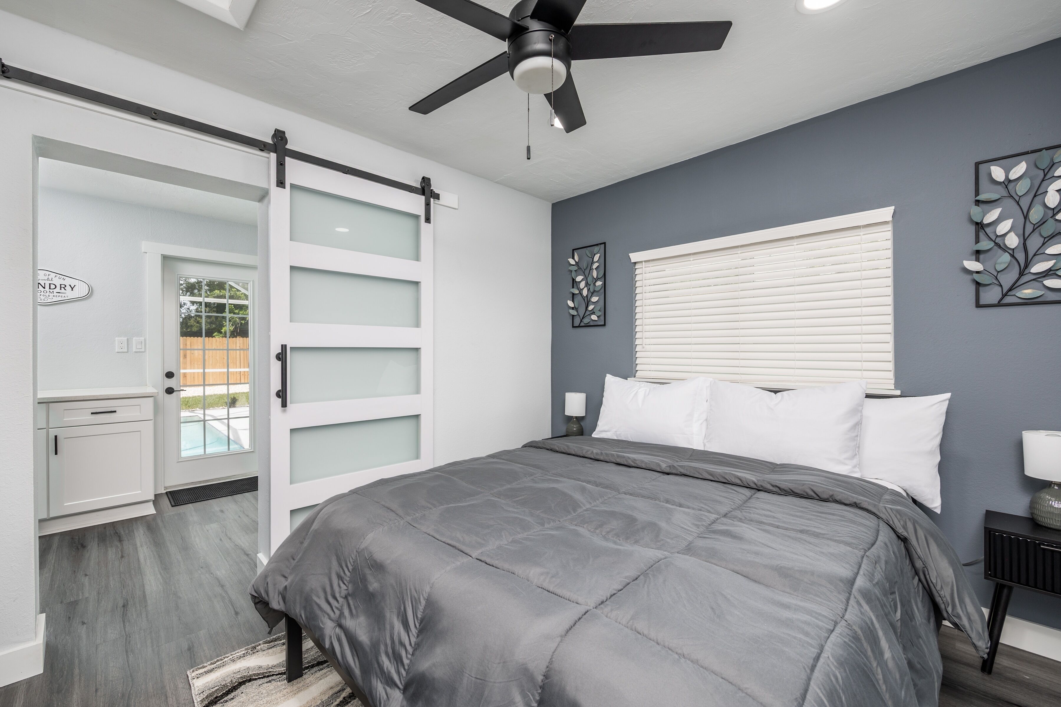 A bedroom with a bed, a ceiling fan, a window with blinds, a door with frosted glass panels, and a wall-mounted artwork.
