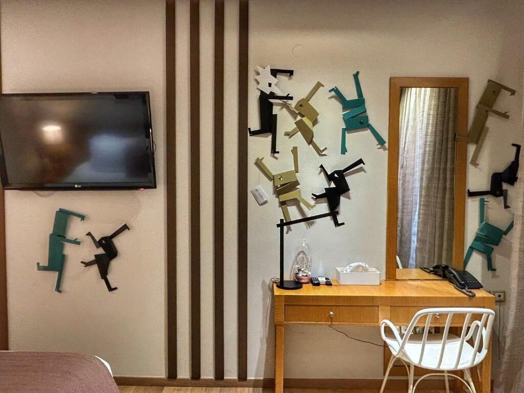 Habitación de hotel con escritorio de madera, una silla blanca, televisor de pantalla plana montado en la pared y arte abstracto en la pared.
