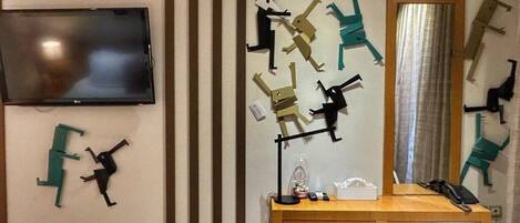 Habitación de hotel con escritorio de madera, una silla blanca, televisor de pantalla plana montado en la pared y arte abstracto en la pared.
